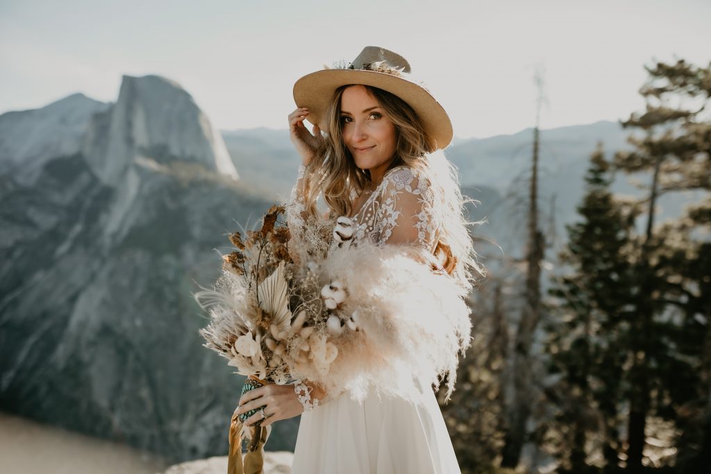 Bride with hand on hat at Glacier Point in Yosemite with Half Dome in the distance.  Be fearless in pursuit of your dreams